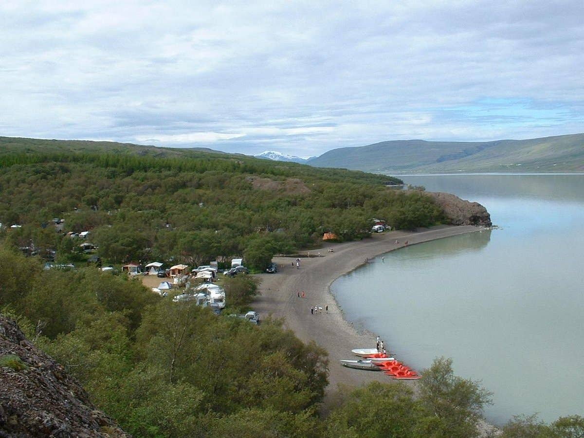 Atlavík shoreline with small boats, parked campers, birch trees, and calm Lake Lagarfljót water.