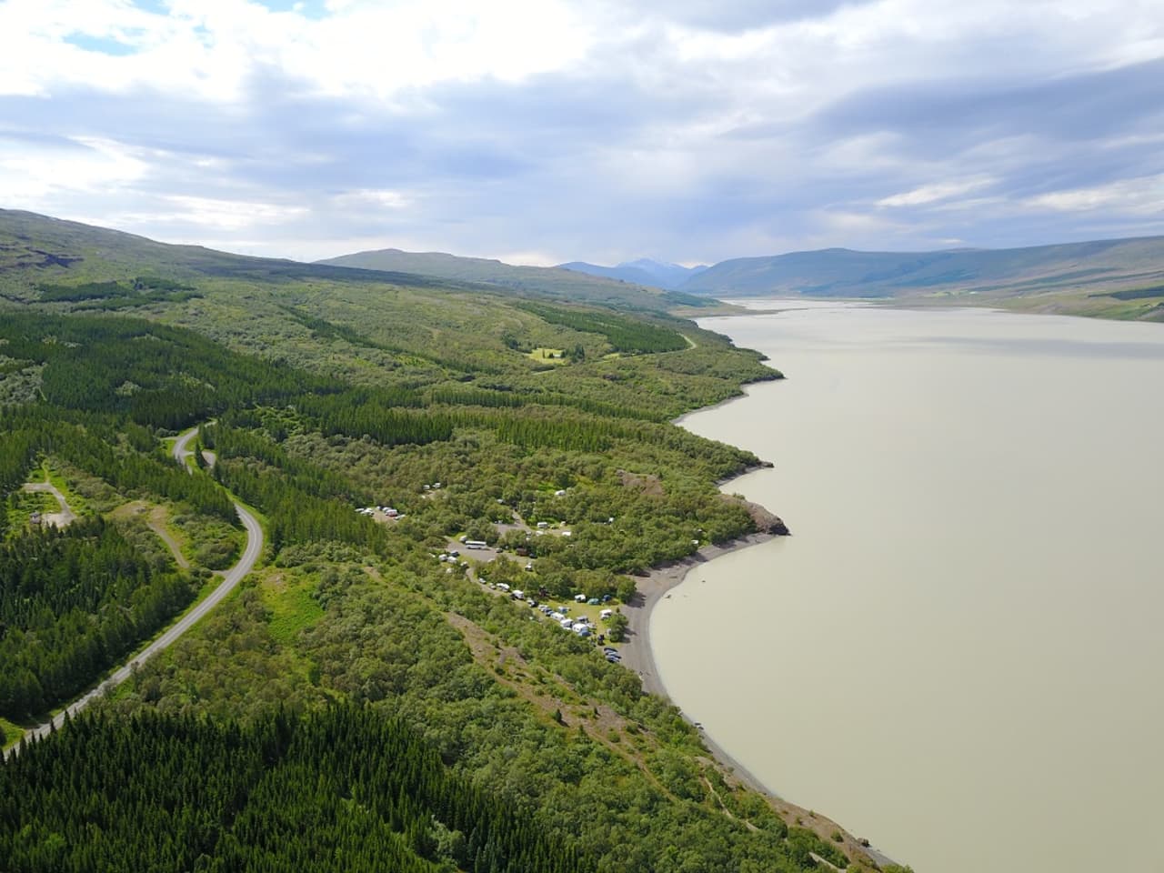 Atlavík cove on Lake Lagarfljót with forest, campsites, a shoreline road, and green East Iceland hills.