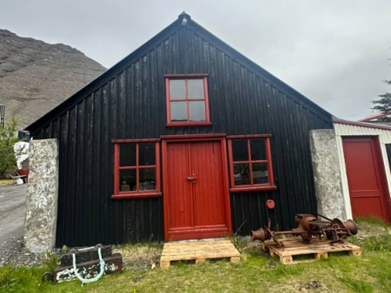 The black timber Old Blacksmith Shop in Bíldudalur with red trim and old tools outside.