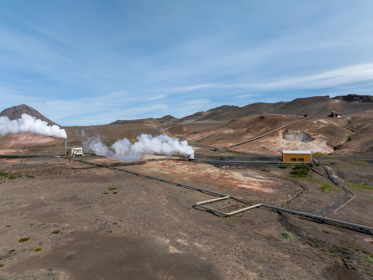 Wide view of Bjarnarflag geothermal pipes, steam vents, yellow station building, and bare volcanic slopes.