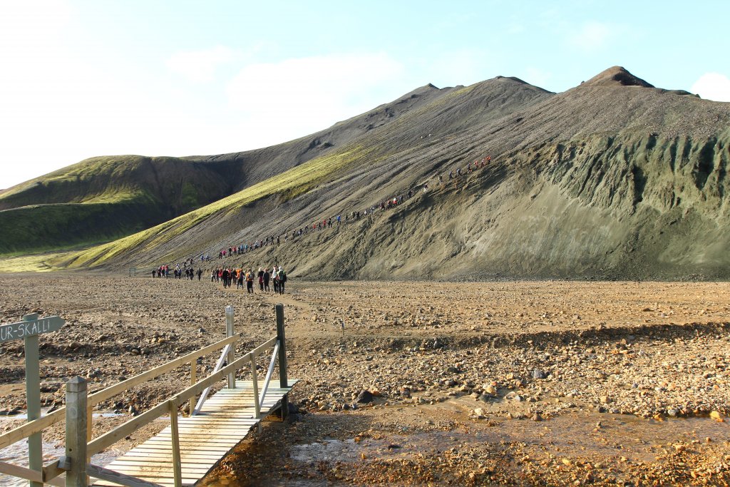 Hikers crossing the bridge from Landmannalaugar toward the blue-black slope of Bláhnjúkur.