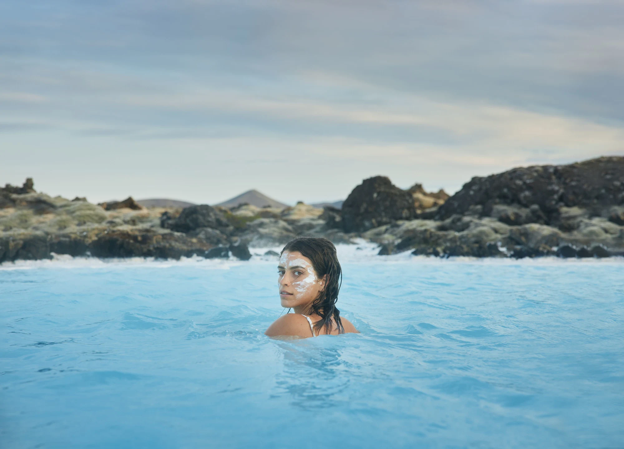 A bather with a silica mask in Blue Lagoon water with black Reykjanes lava behind her.