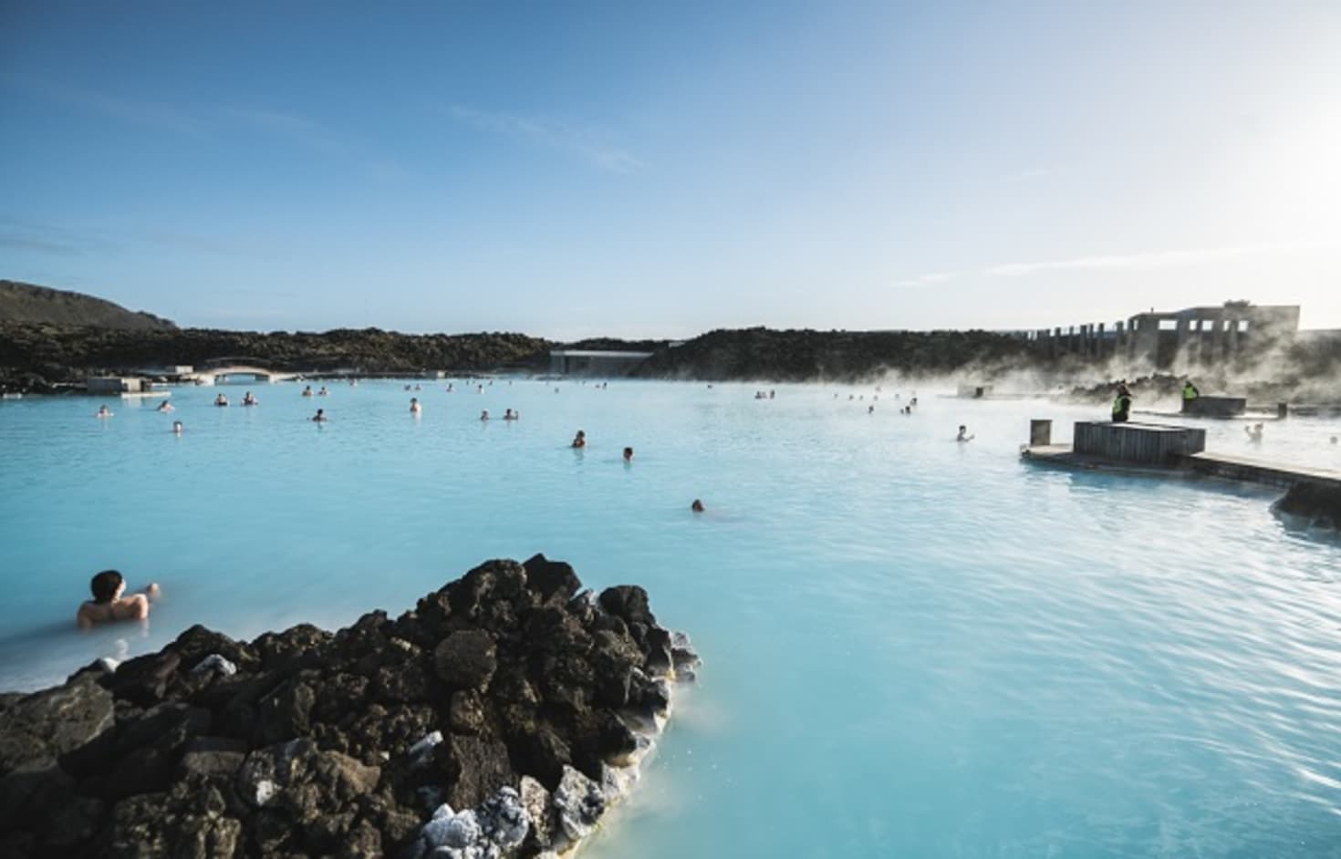 Visitors spread across the Blue Lagoon with steam, black lava, and spa buildings around the water.