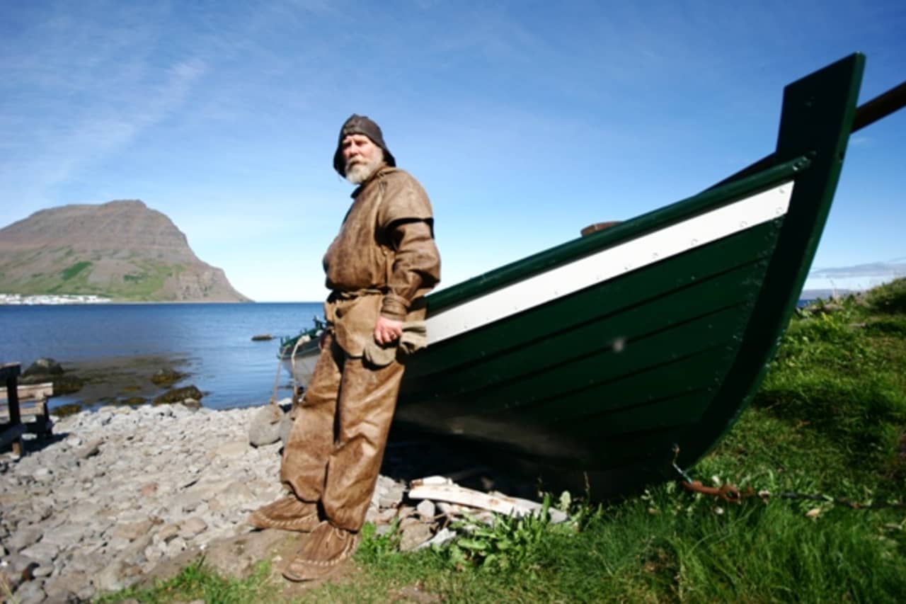 Guide in traditional fishing clothing beside a wooden boat at Ósvör near Bolungarvík.