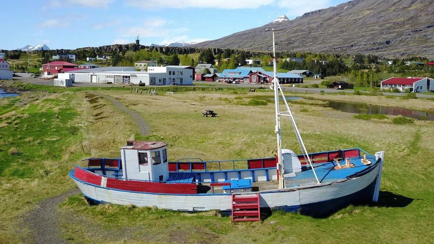 A small boat on shore in Breiðdalsvík with village buildings and steep Eastfjords mountains behind.