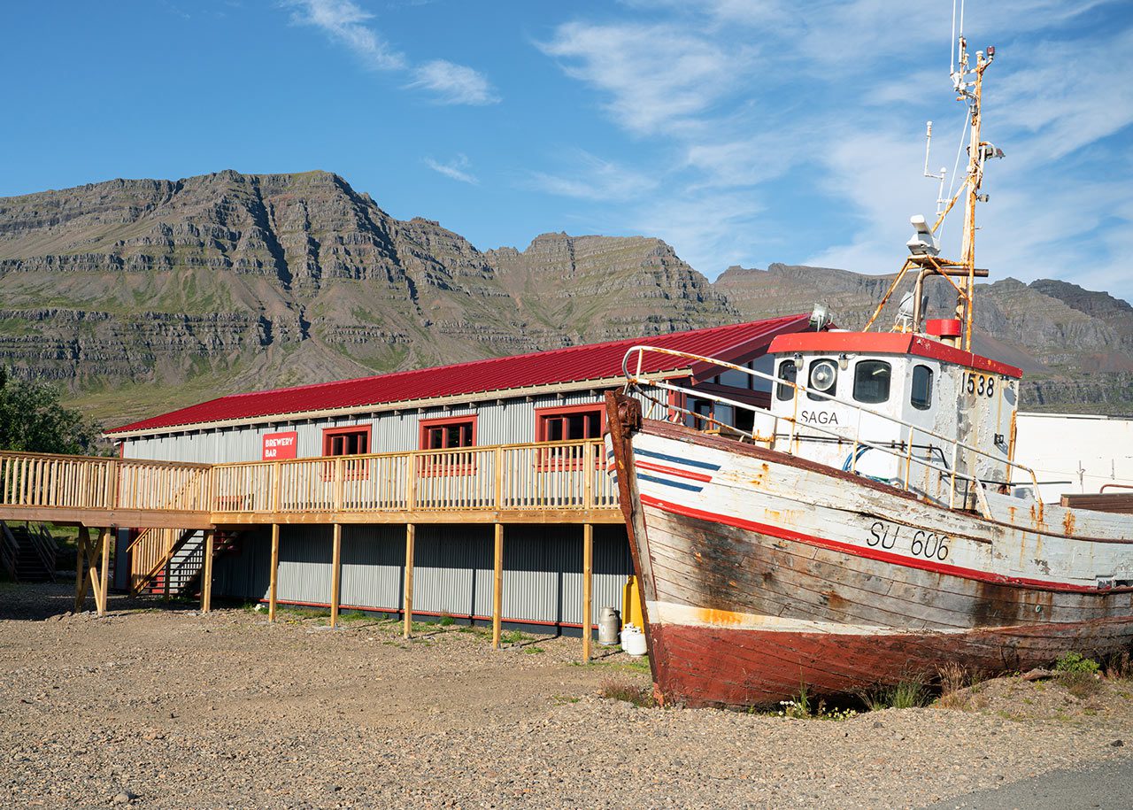 A weathered boat and low local buildings in Breiðdalsvík below layered Eastfjords mountains.