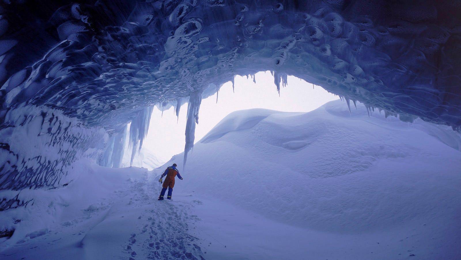 Person in technical outerwear inside a blue ice cave at Brúarjökull with snow and icicles around the entrance.