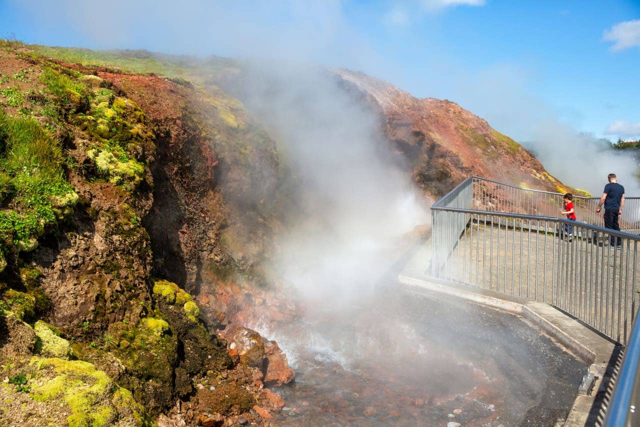 Visitors standing behind a metal viewing platform beside steam and red-brown rock at Deildartunguhver.