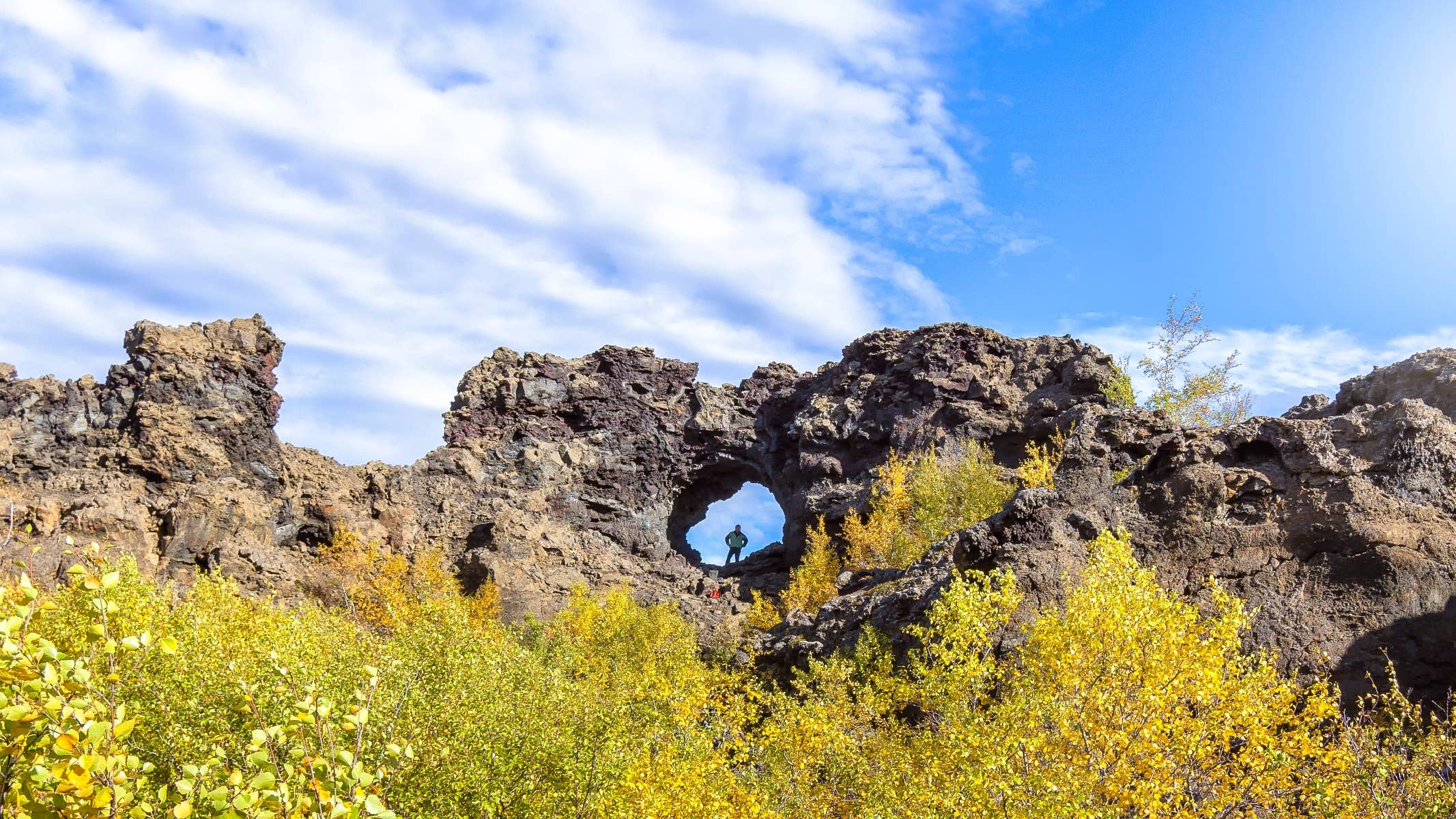 Wide Dimmuborgir lava wall with a small natural arch and yellow autumn birch below it.