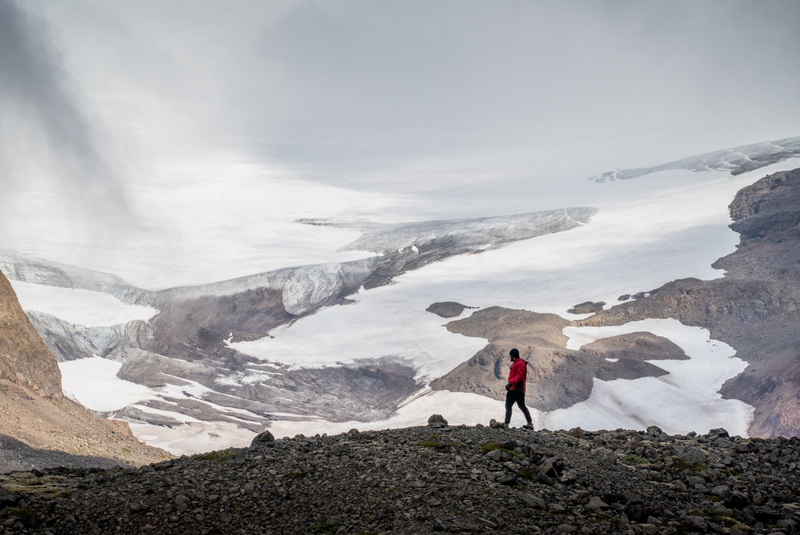 A hiker standing on dark gravel above snowy Drangajökull ice near Kaldalón in the Westfjords.