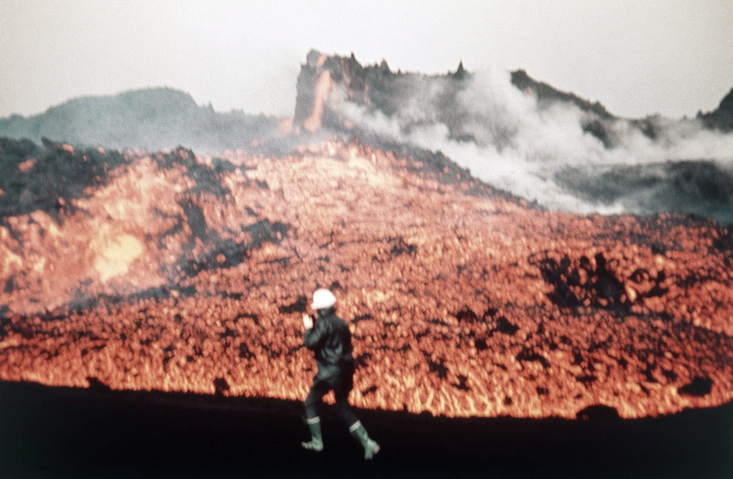 A rescue worker walking near glowing lava and smoke during the 1973 Eldfell eruption on Heimaey.