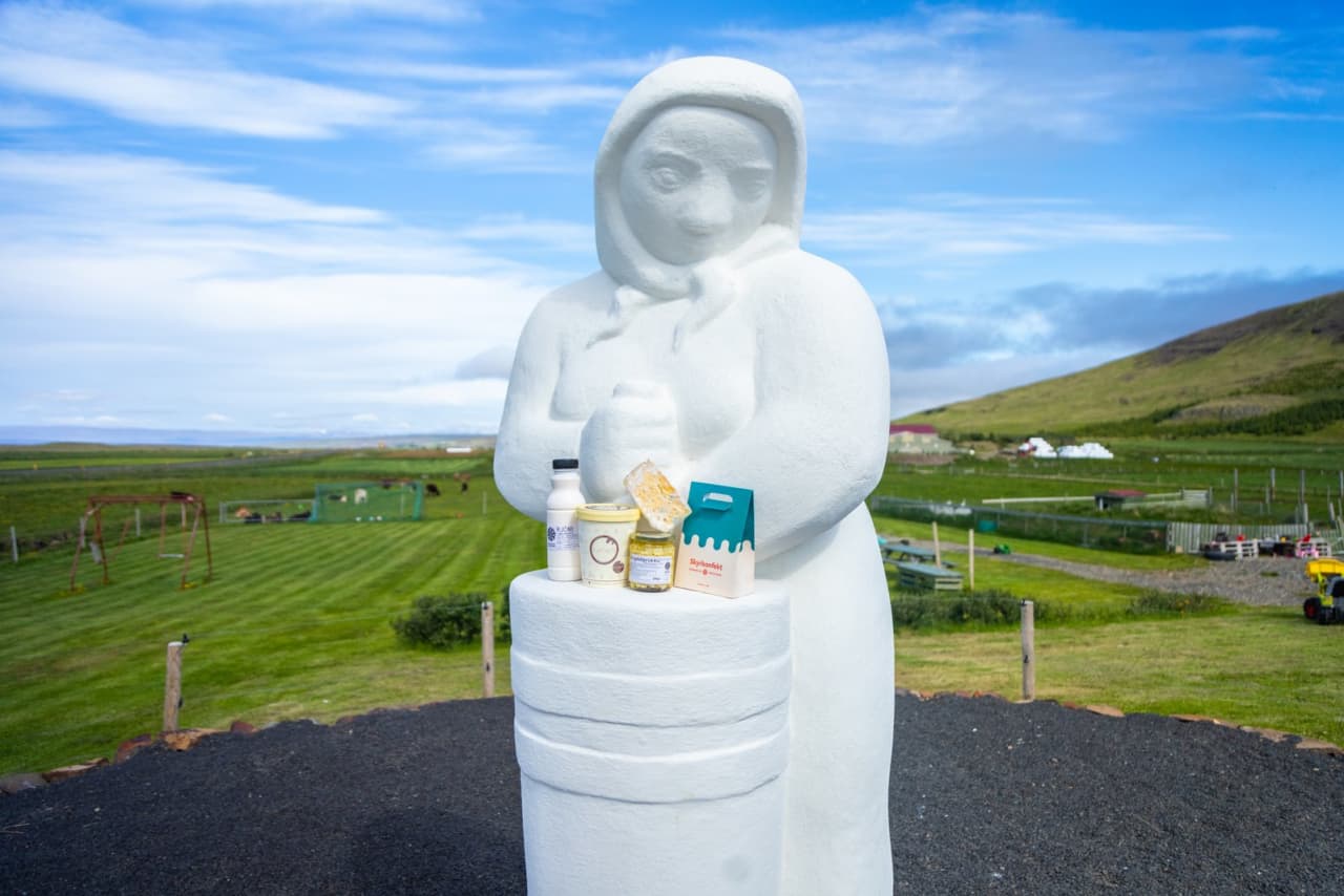 A white creamery statue at Erpsstaðir with farm fields and products behind it.