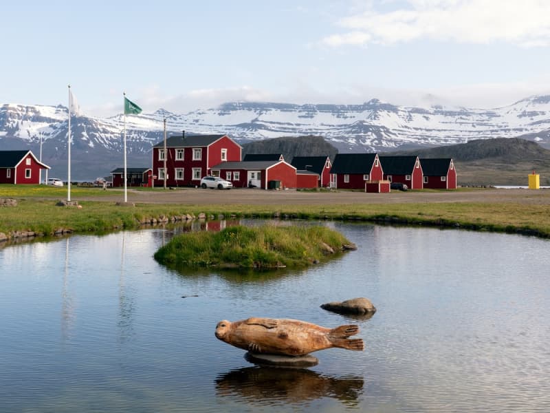 Red buildings at Mjóeyri reflected in a pond with snowy mountains behind Eskifjörður.