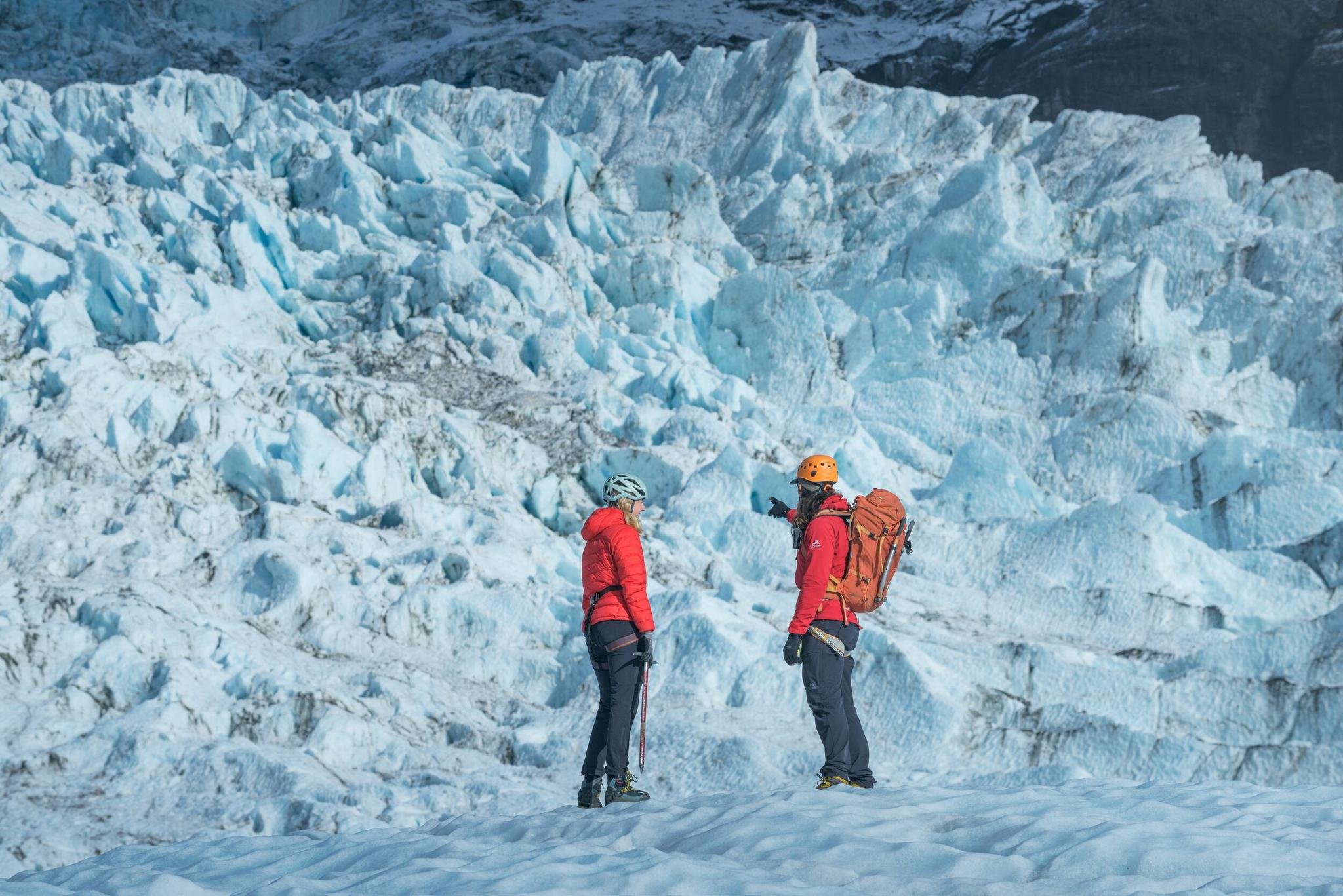 Guide and visitor in helmets standing below broken blue ice on Falljökull Glacier.