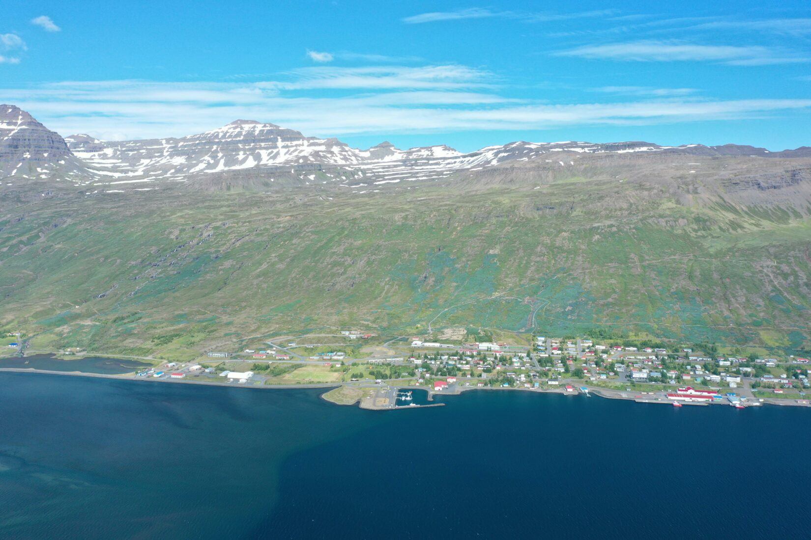 Aerial view of Fáskrúðsfjörður village, harbor, fjord water, and steep Eastfjords mountains.
