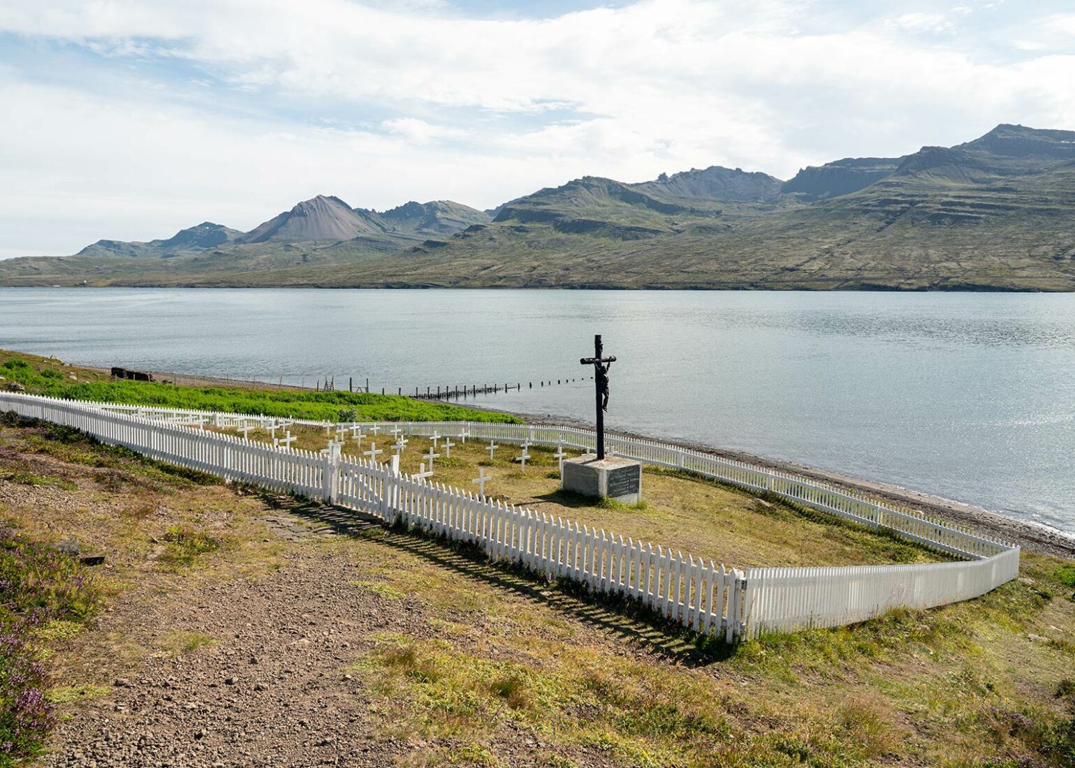 French cemetery memorial near Fáskrúðsfjörður with stone wall, grass, and mountains in the distance.
