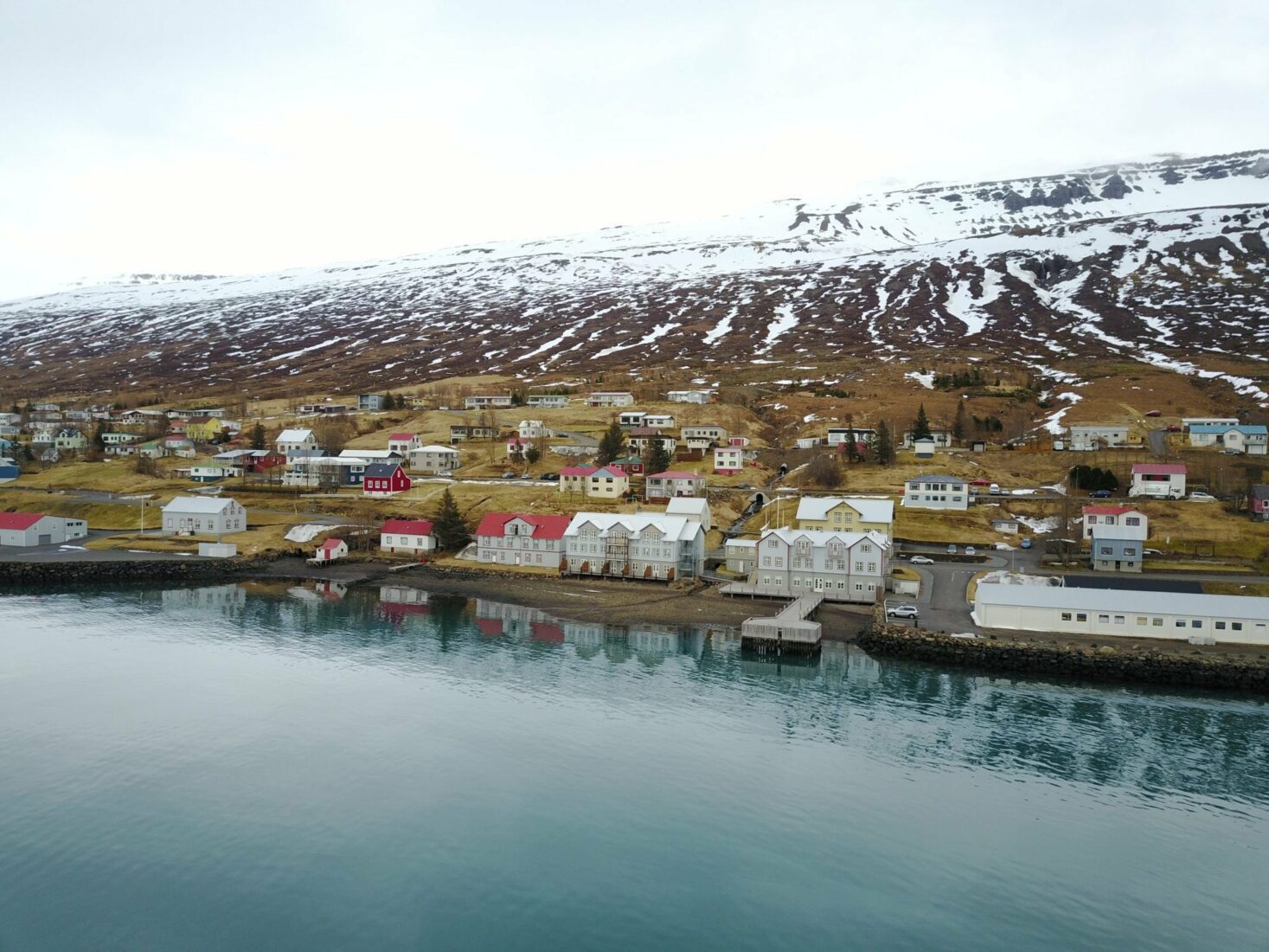 French heritage buildings by the water in Fáskrúðsfjörður with snowy Eastfjords slopes behind.