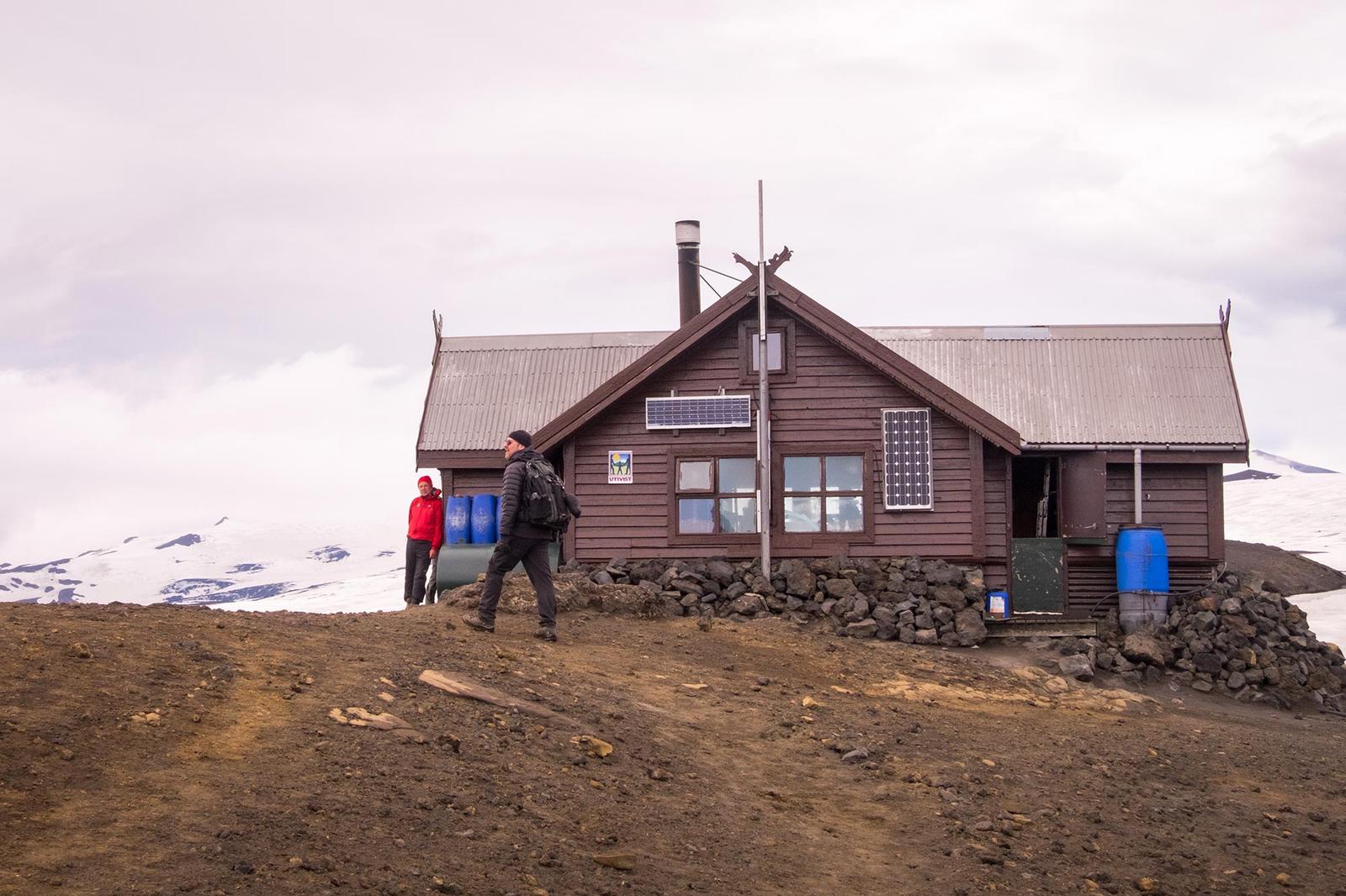 A small mountain hut on Fimmvörðuháls with hikers and snowfields around it.