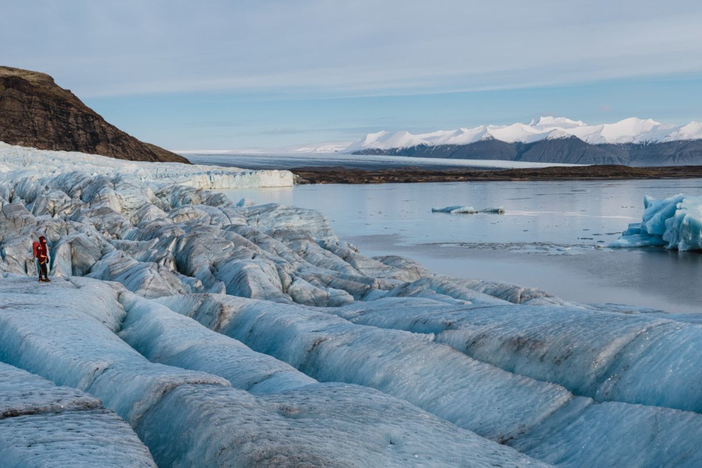 A guided visitor standing on blue glacier ice above Fjallsárlón with mountains in the distance.