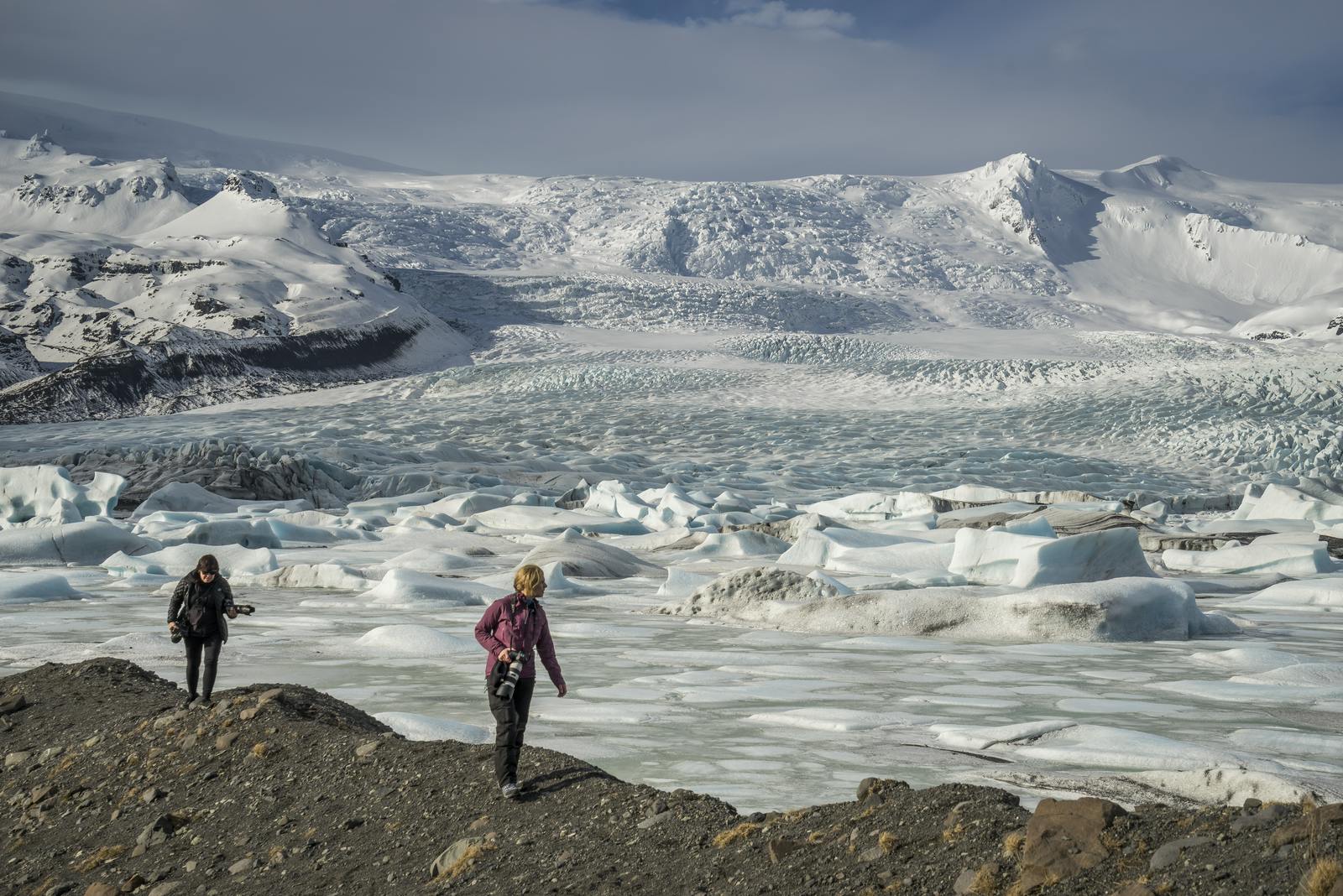 Travelers standing near Fjallsárlón with Fjallsjökull glacier rising behind floating ice.
