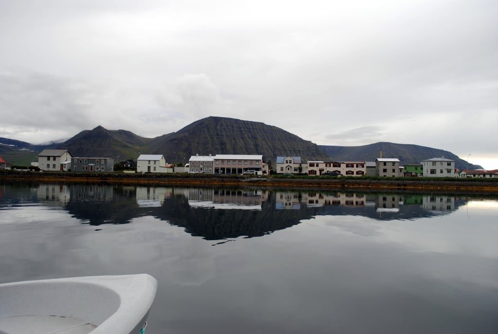 Flateyri houses reflected in calm harbor water below dark Westfjords mountains.