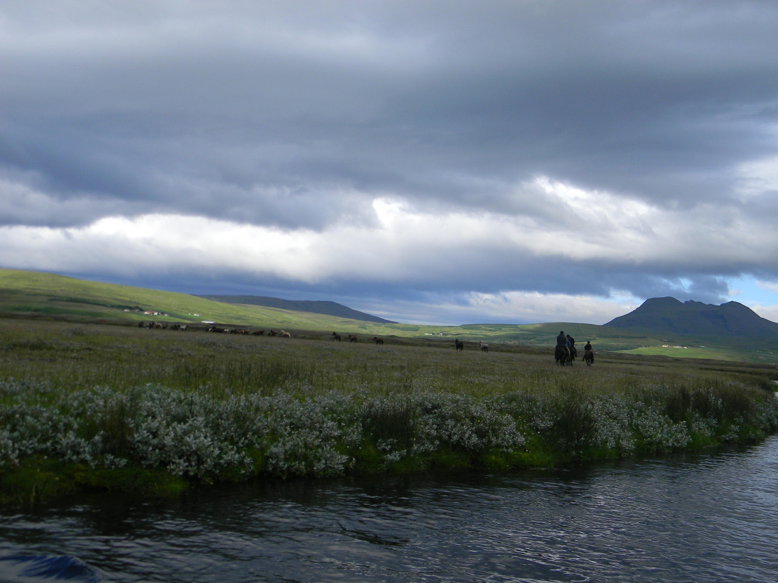 Riders crossing open Fljótshlíð countryside below dark clouds and low mountains.