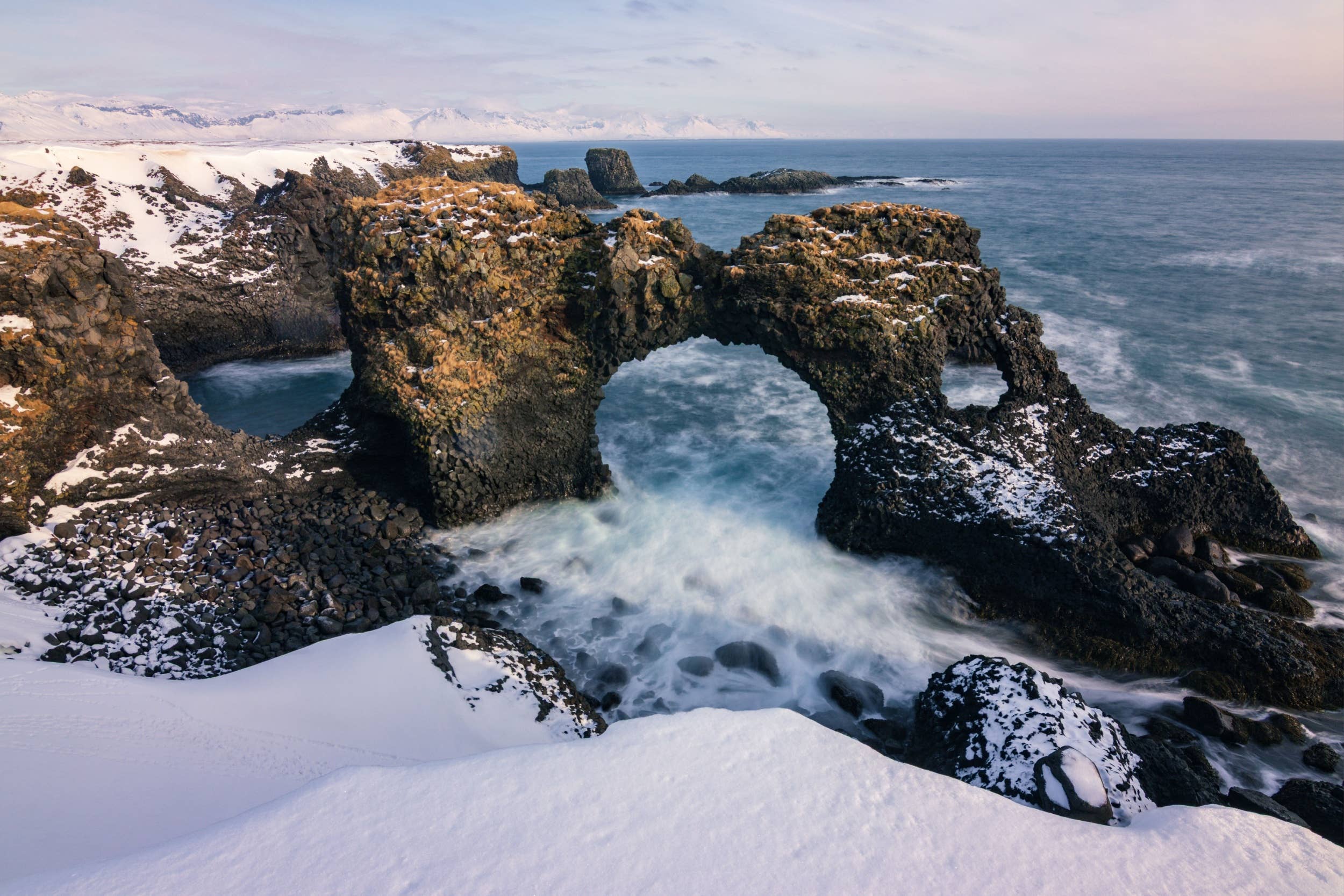 Snow around Gatklettur sea arch with waves surging through the basalt opening.