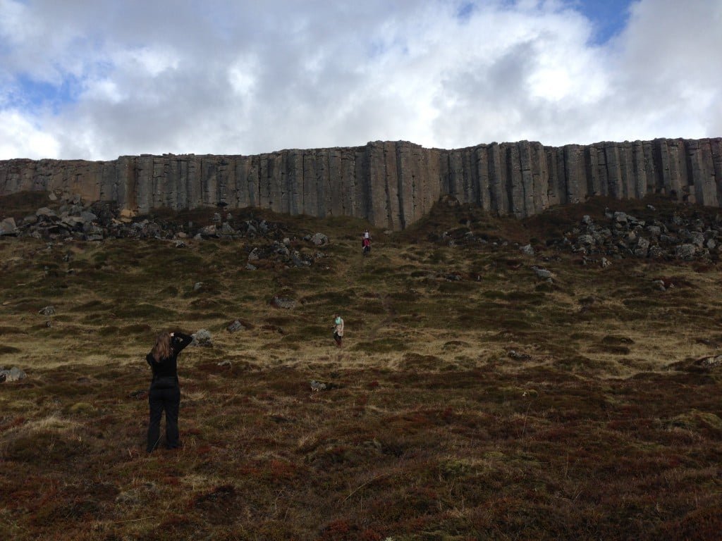 People walking below the Gerðuberg basalt-column wall on a grassy uneven slope.