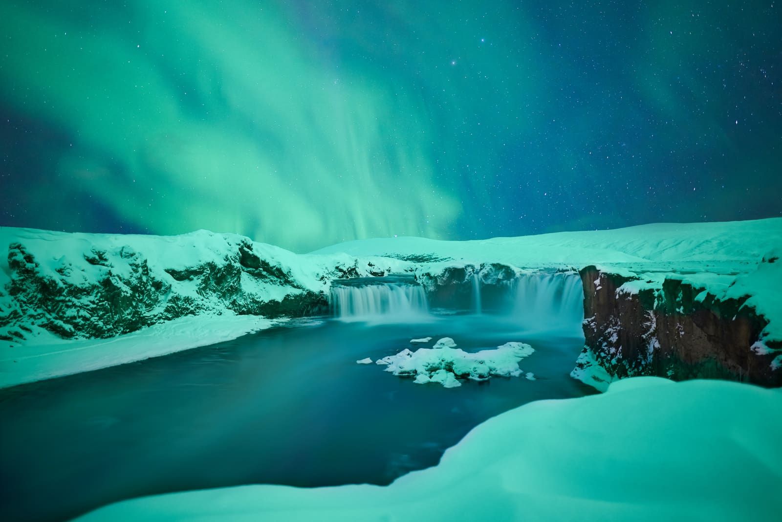 Northern lights above snowy Goðafoss with part of the waterfall visible below the winter sky.