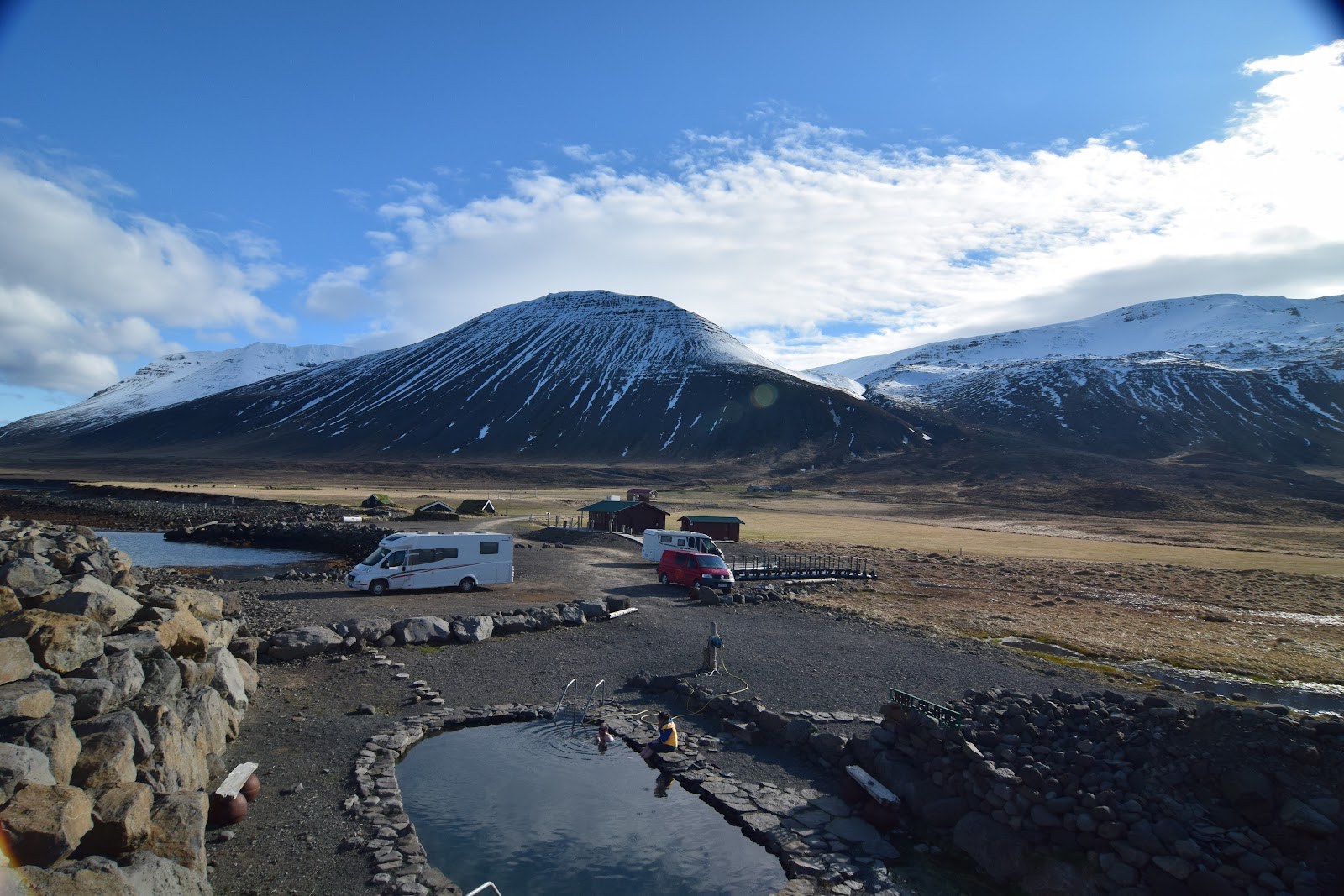 Grettislaug pools, parked vehicles, gravel paths, and snow-streaked mountains in the Reykjaströnd landscape.