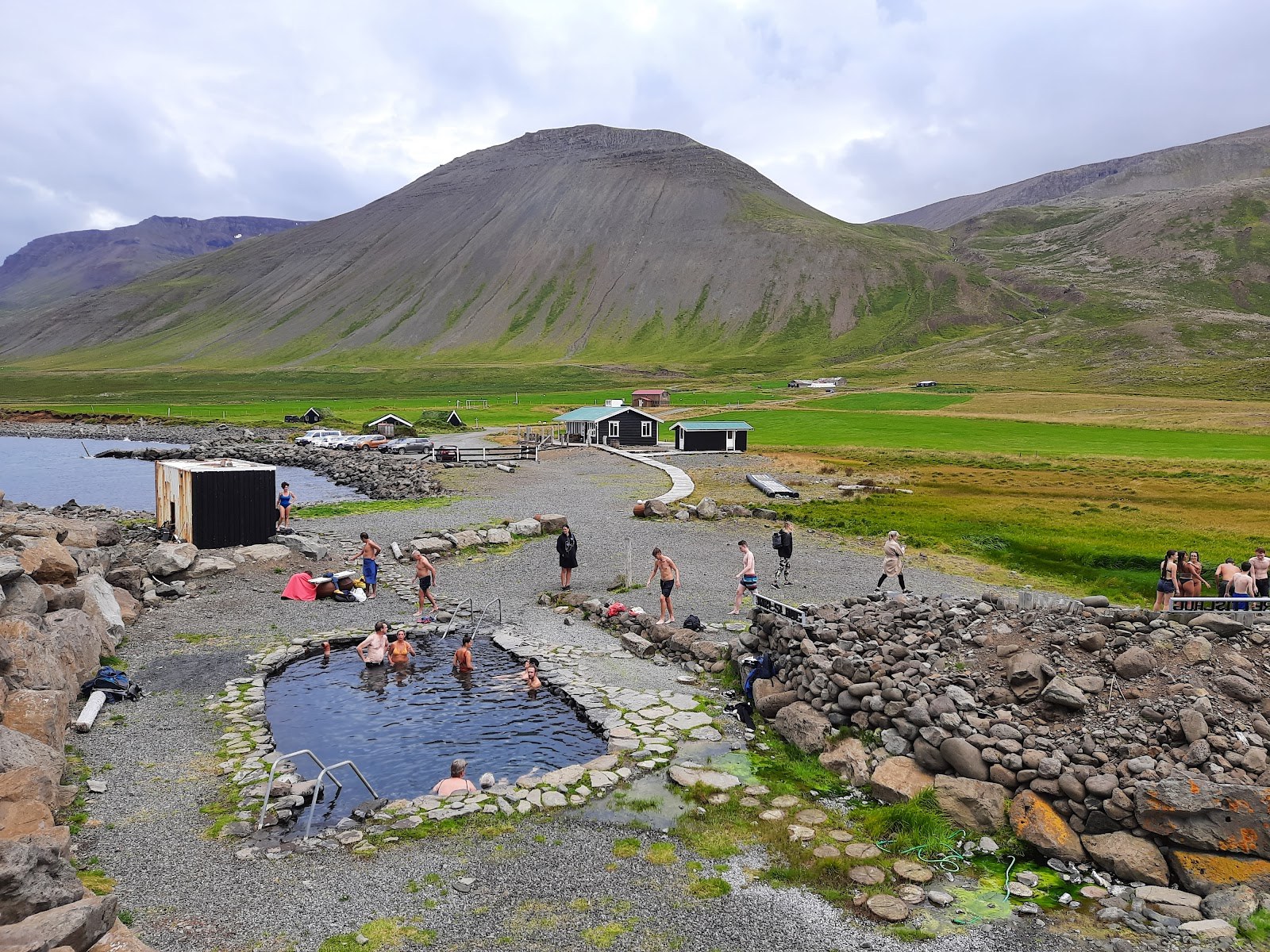 People soaking in Grettislaug with gravel paths, low buildings, and steep Skagafjörður mountains behind the pool.