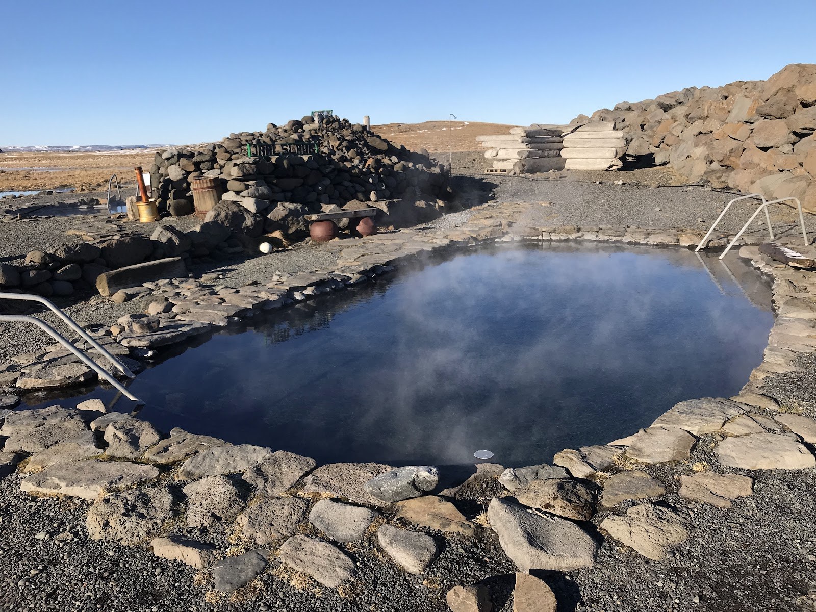 Steam rising from a stone-lined pool at Grettislaug with low rock walls and gravel around the bathing area.