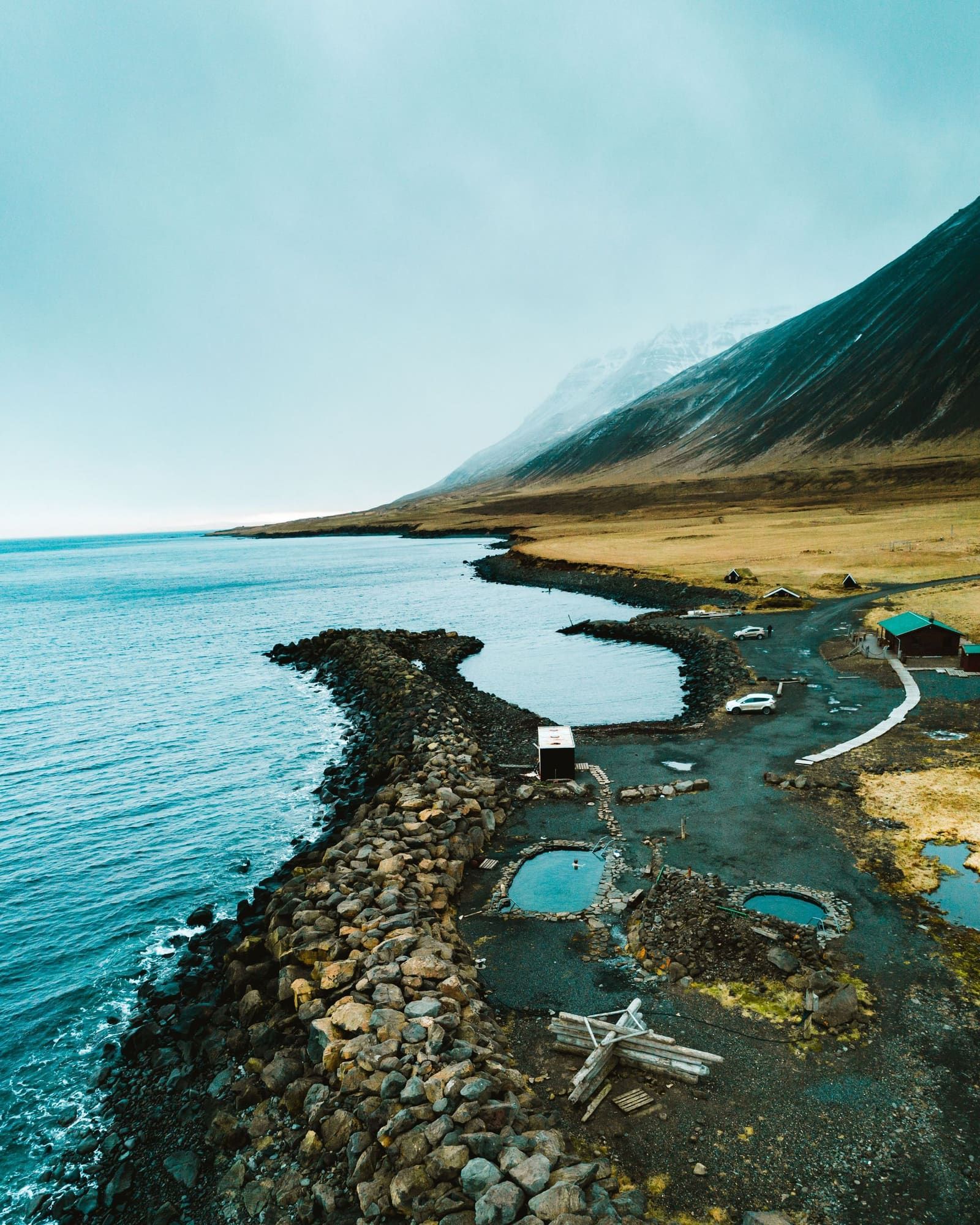 Coastal overview of Grettislaug beside the sea at Reykjaströnd with pools, paths, rocks, and mountains.