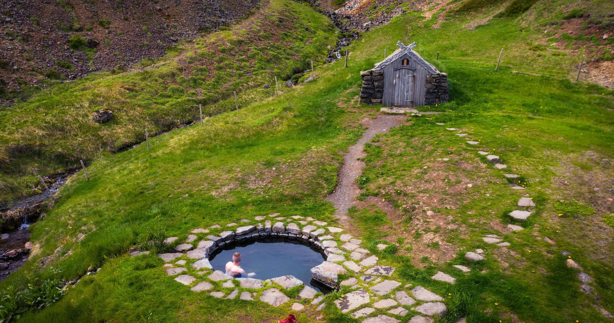 Overhead view of Guðrúnarlaug pool, footpath, turf hut, and grassy Sælingsdalur slope.