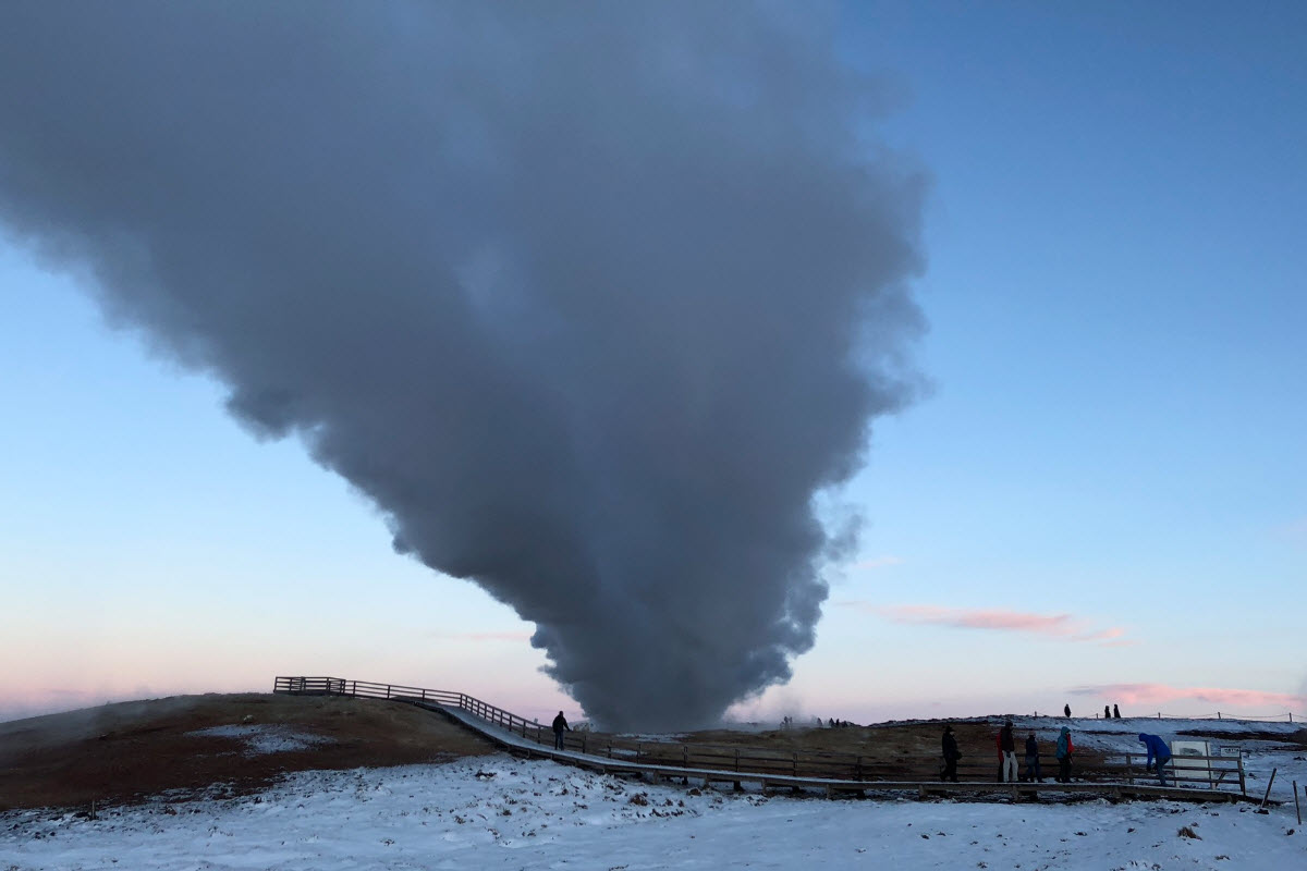 Visitors walking on a snowy boardwalk beside a dense steam plume at Gunnuhver.