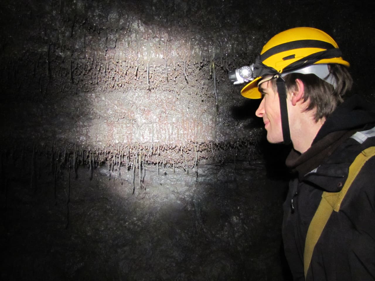 Visitor wearing a helmet and headlamp beside a dark lava wall inside a Hallmundarhraun cave.