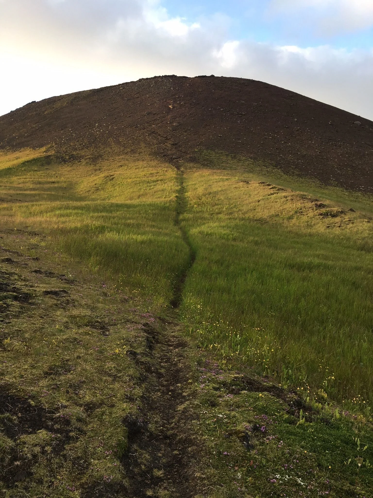 A narrow grassy path climbs the dark upper slope of Helgafell.