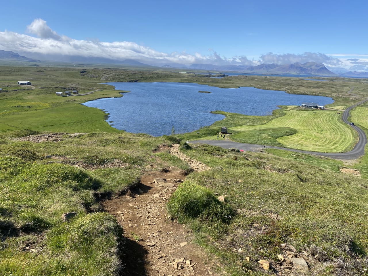 A rocky path descends Helgafell toward a blue lake, farms, and distant Snæfellsnes mountains.