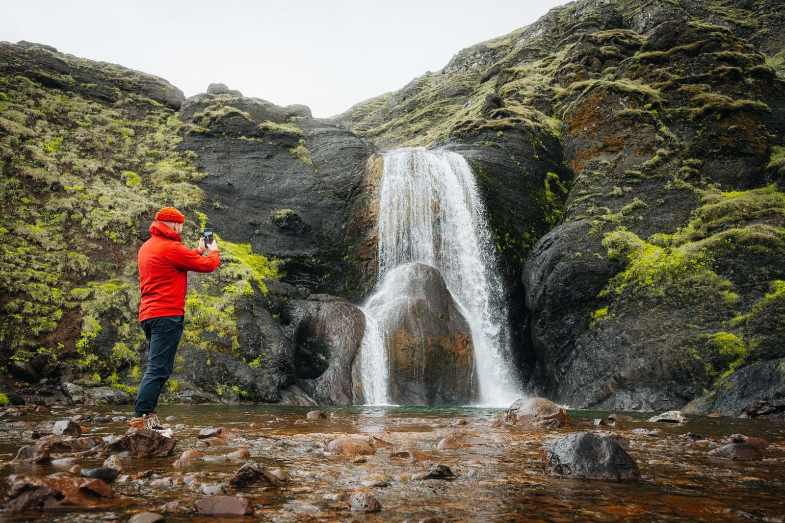 Visitor in a red jacket facing Helgufoss waterfall in the dark ravine at Mosfellsdalur.