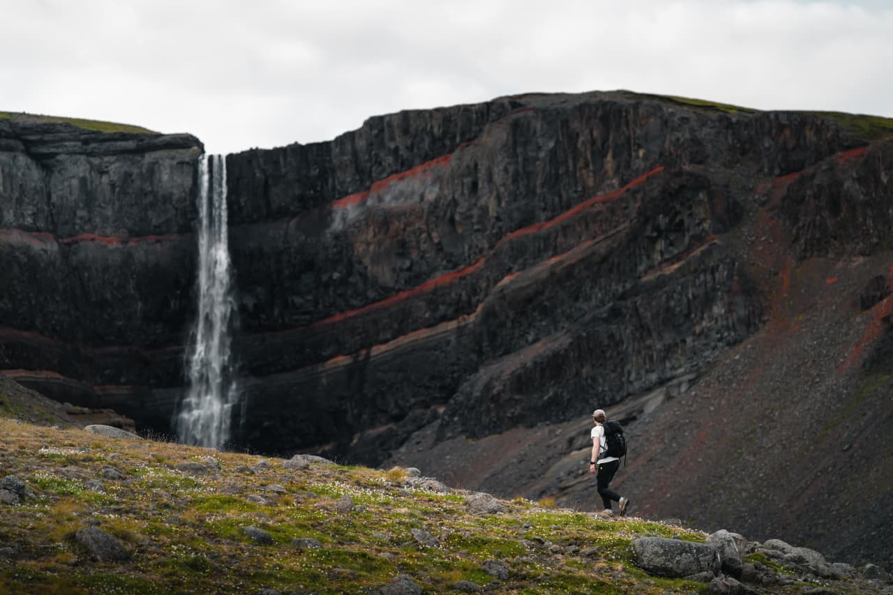 A hiker on the Hengifoss trail approaching the tall waterfall and red-striped basalt gorge.