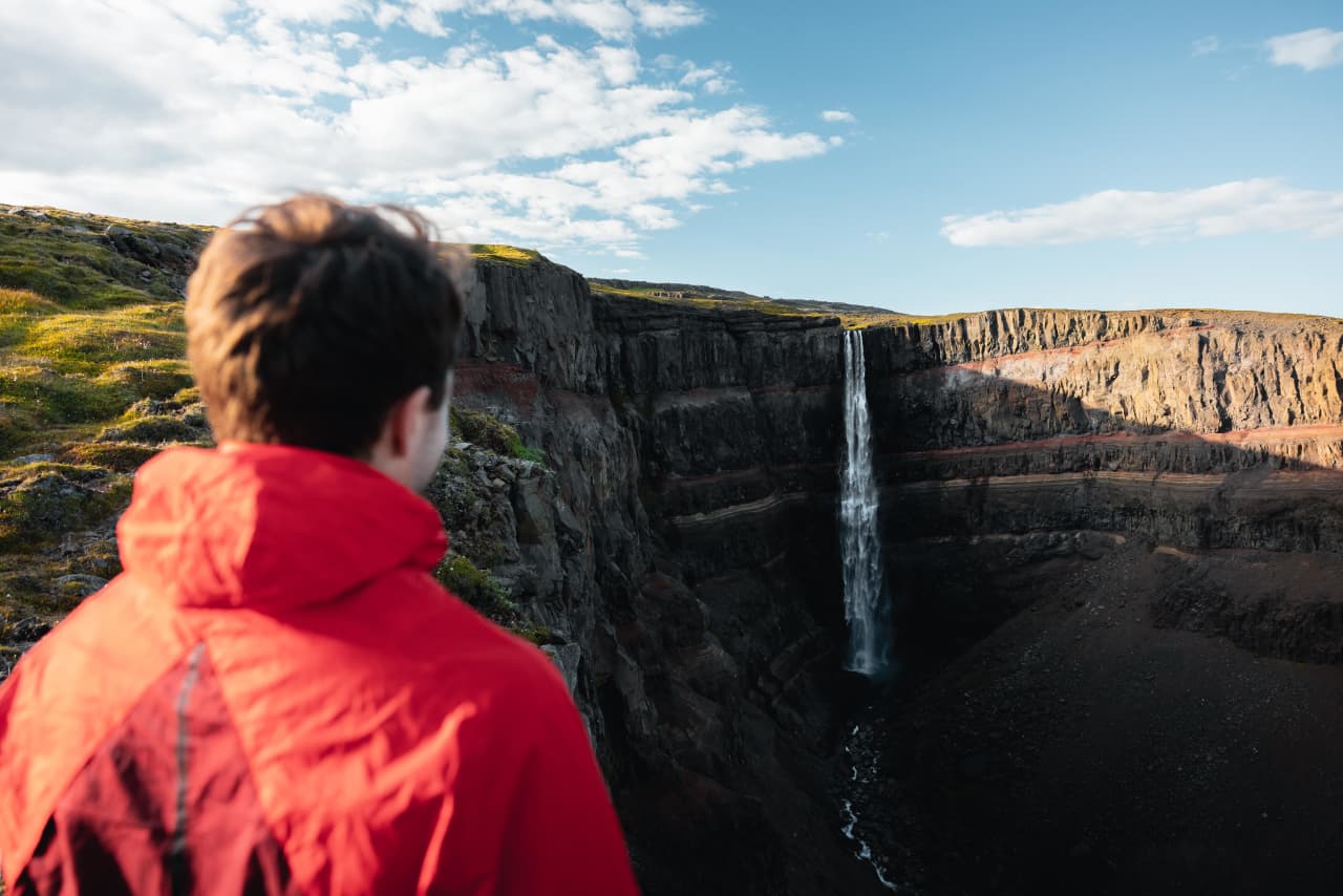 A person in a red jacket looking across the Hengifoss gorge toward the waterfall and red sediment bands.