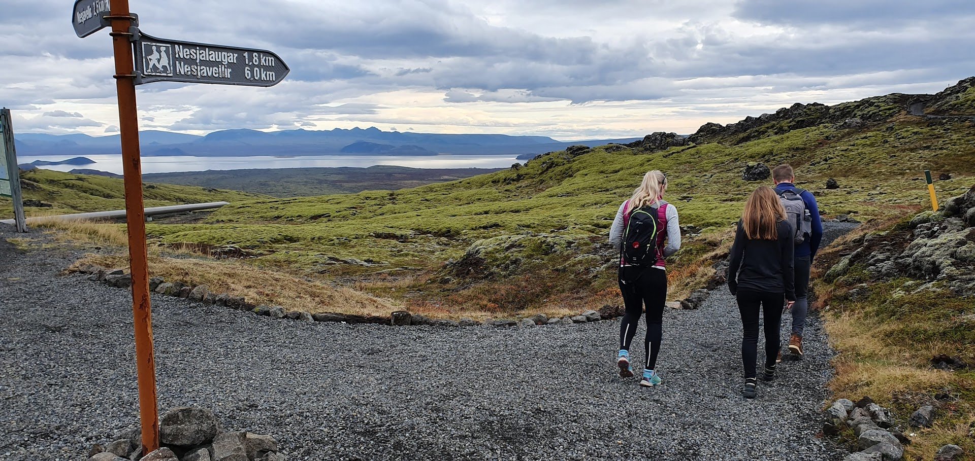 Walkers following a marked Hengill trail with a signpost, lava slopes, and Þingvallavatn in the distance.