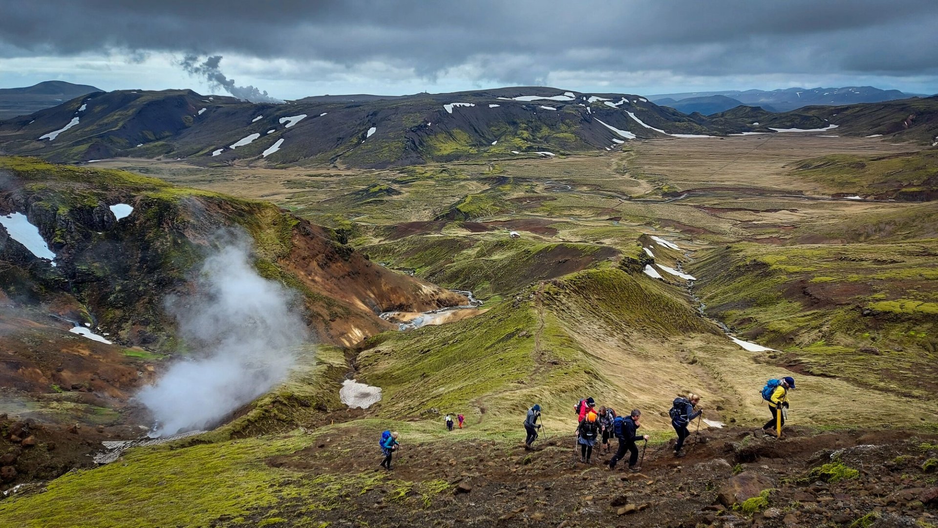 Hikers on a ridge in the Hengill geothermal landscape above broad valleys and steam.