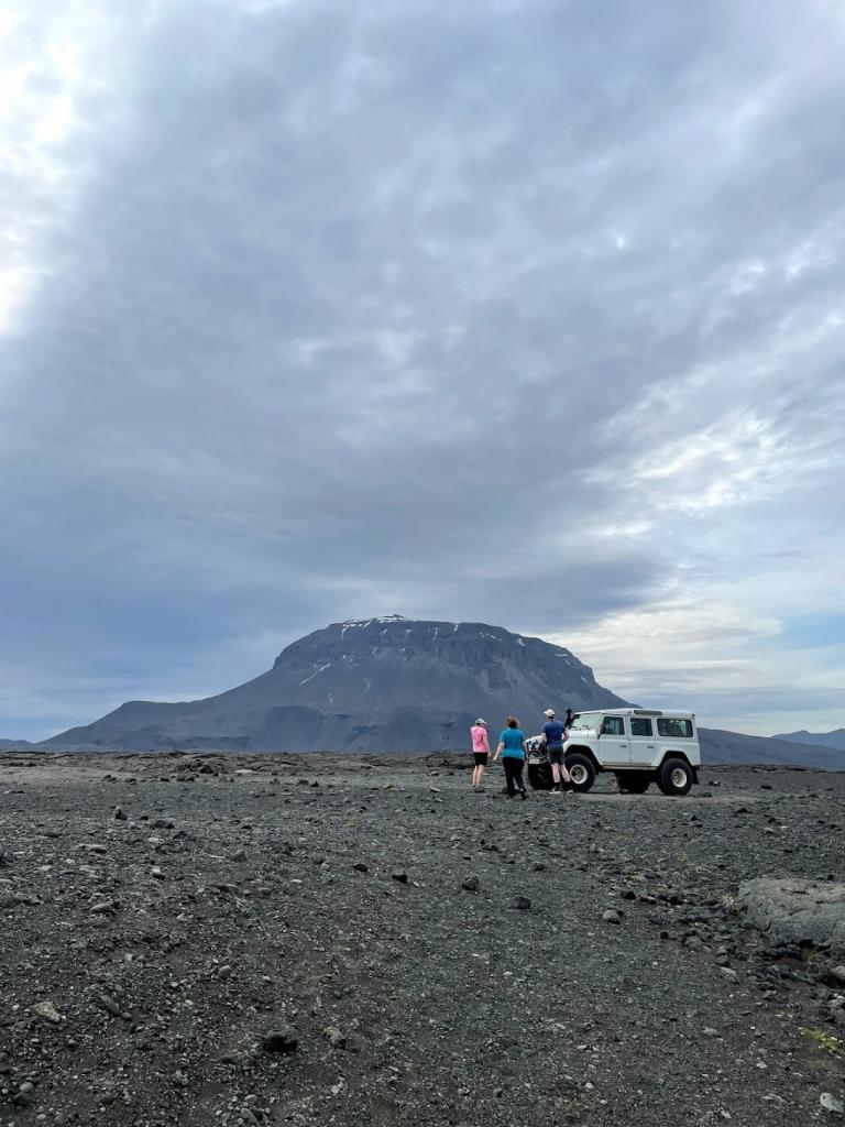 A highland vehicle and travelers standing on dark lava ground below Herðubreið.
