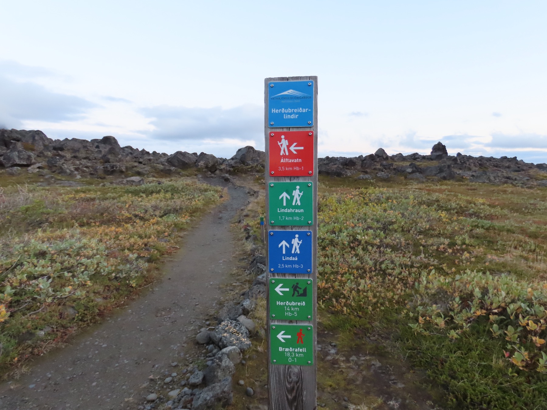 A hiking sign at Herðubreiðarlindir pointing across low vegetation and lava in the highlands.