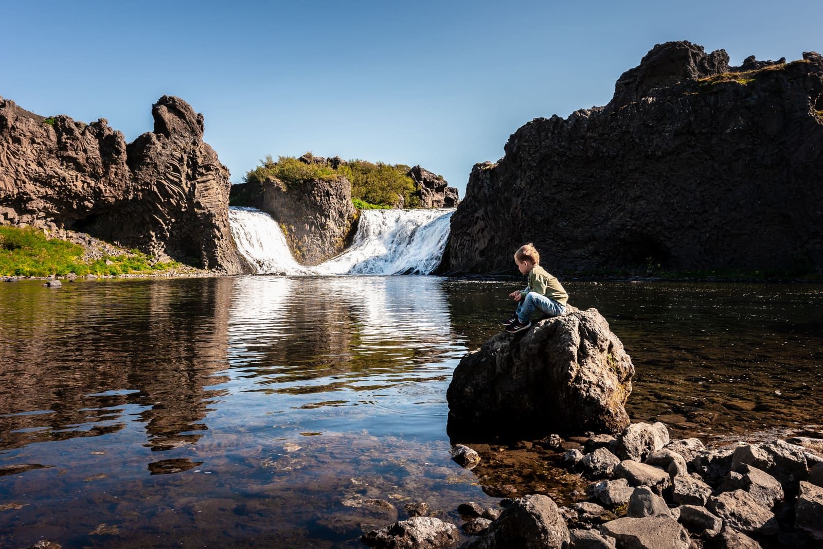 Hjálparfoss waterfall flowing between dark basalt formations into a calm river pool.