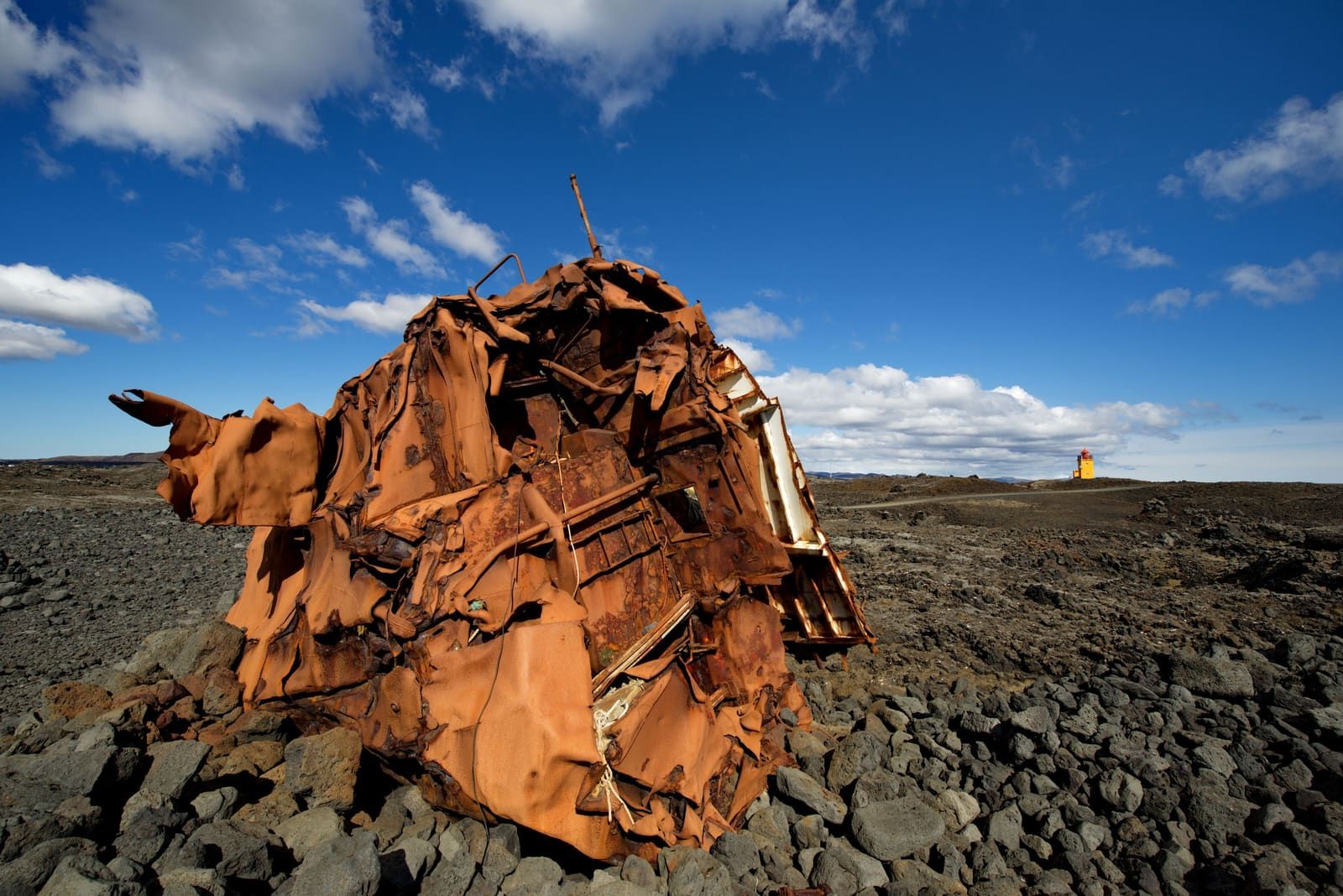 Rusting shipwreck remains on black lava at Hópsnes with the orange Hópsnesviti lighthouse in the distance.