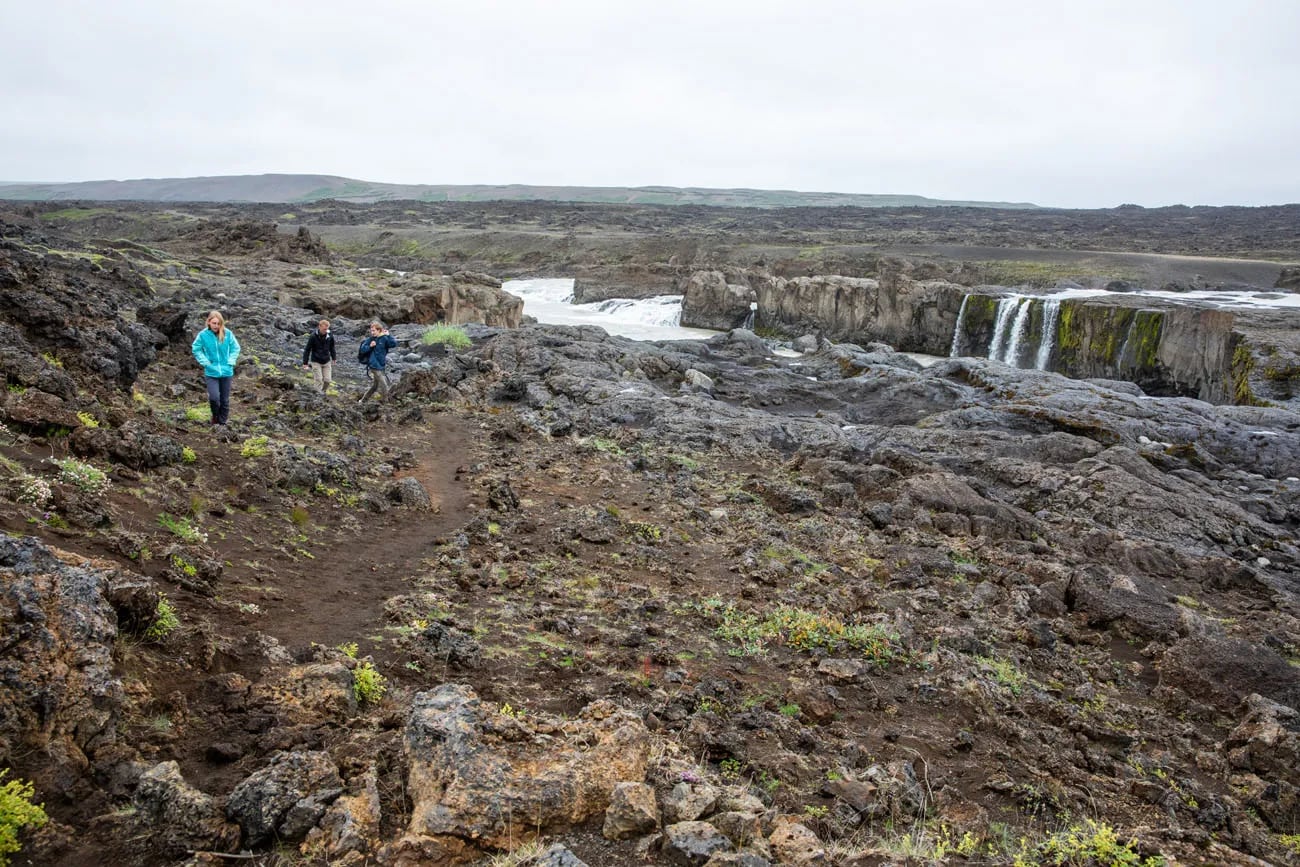 Visitors walking over rough dark ground toward Hrafnabjargafoss with the waterfall in the distance.