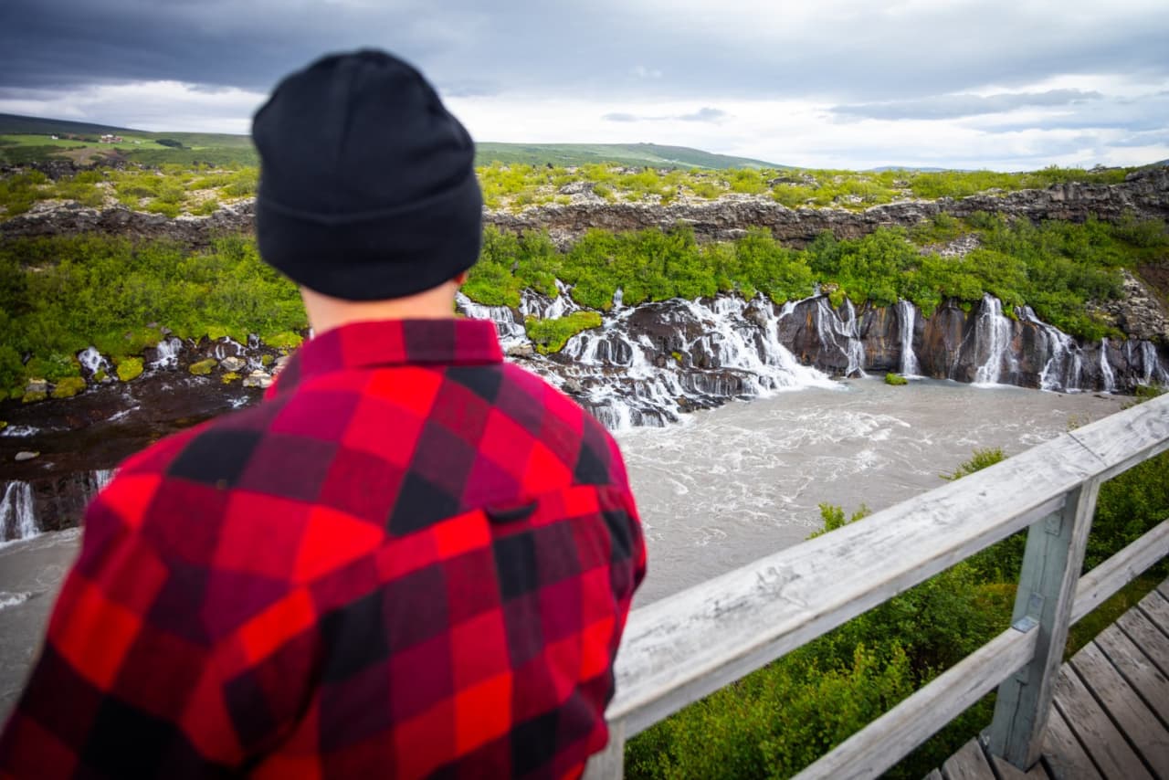 Traveler looking from the Hraunfossar viewing platform toward the lava-fed waterfalls and Hvita river.