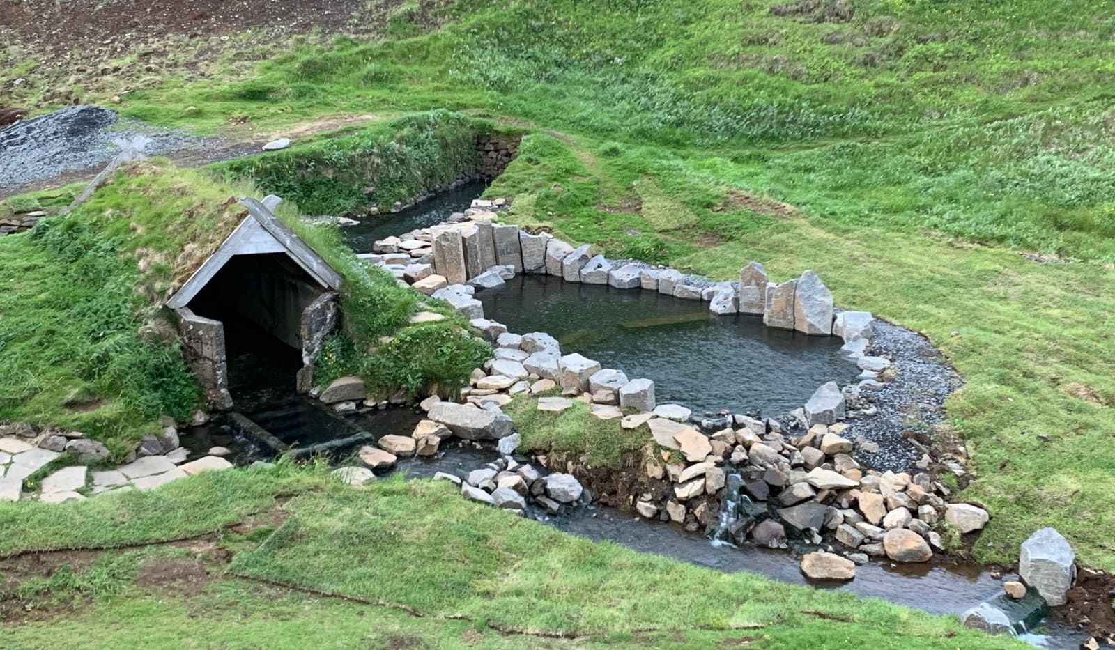 Hrunalaug's main pool and old grass-roofed shelter beneath green hills near Flúðir.